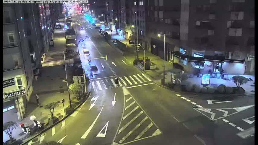 A nighttime, high-angle view of a city street intersection with several cars and pedestrians, some buildings, and streetlights illuminating the scene.