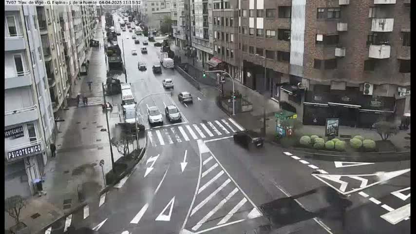 A wet city street intersection shows several cars driving amidst buildings on a rainy day.