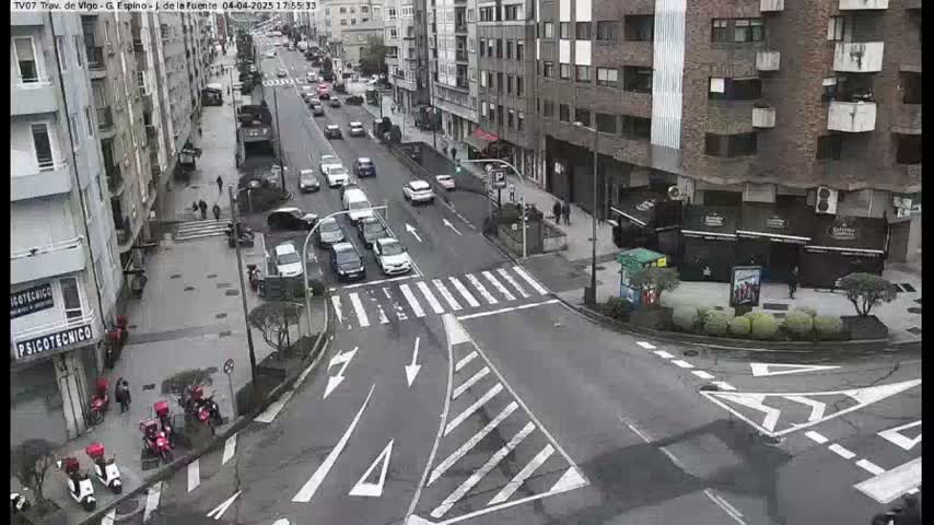 A city intersection with moderate traffic on a cloudy day, several cars are moving along the streets, and there are pedestrians and some scooters at the side of the road.