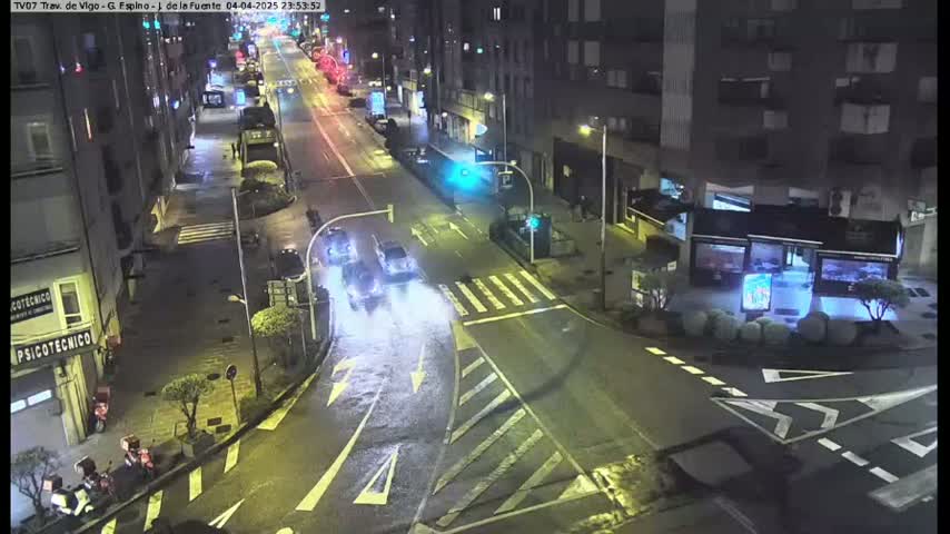 A wet, nighttime street scene shows several cars driving on a multi-lane road that intersects with other roads, surrounded by buildings.