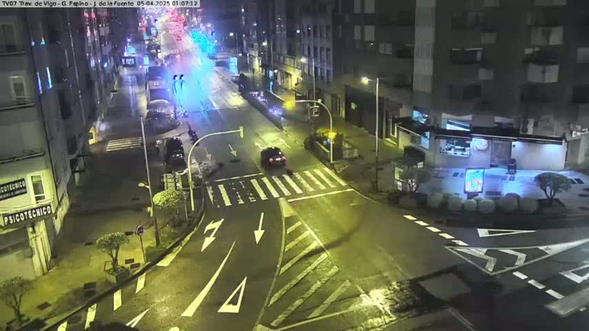 A nighttime street intersection with a few cars and buildings visible under wet street conditions.