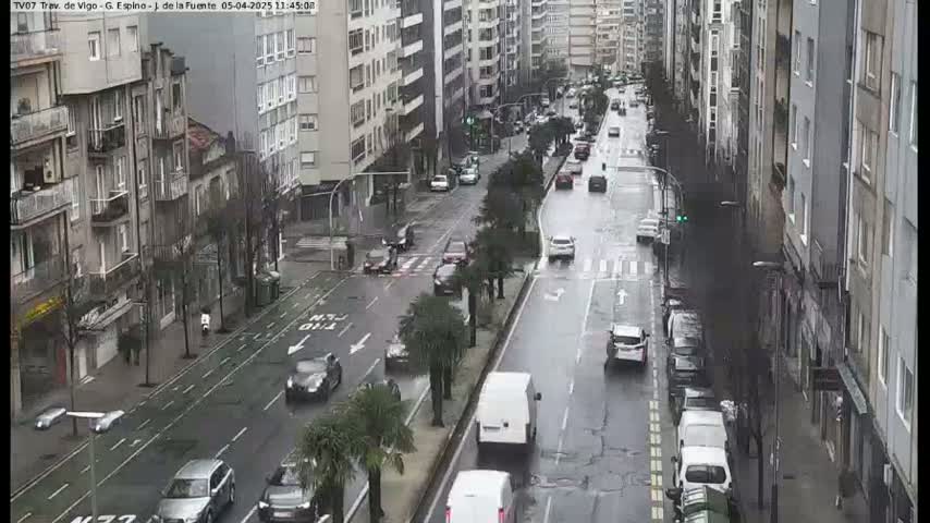 A wet city street scene shows several cars driving past multi-story buildings on a rainy day.