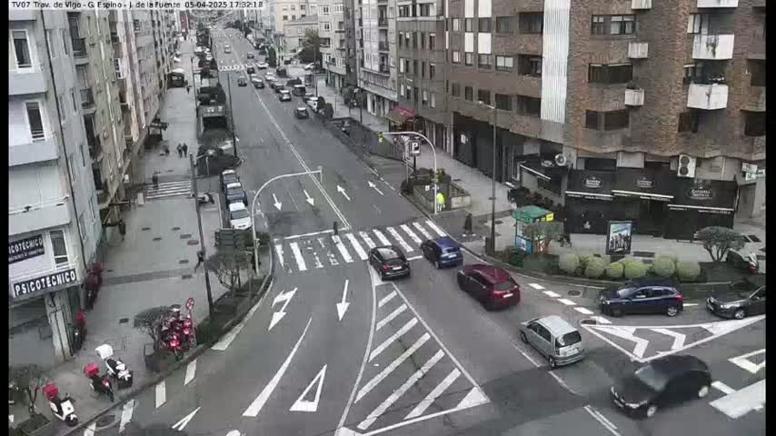 A street scene shows several cars at an intersection in a city with buildings lining both sides of the street on an overcast day.