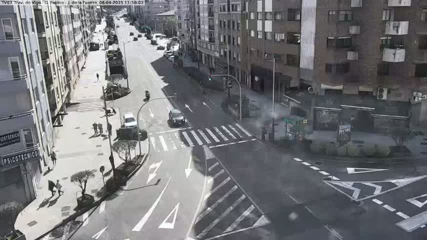 A sunny day reveals a city street intersection with several cars and pedestrians, and buildings lining the sides of the street.