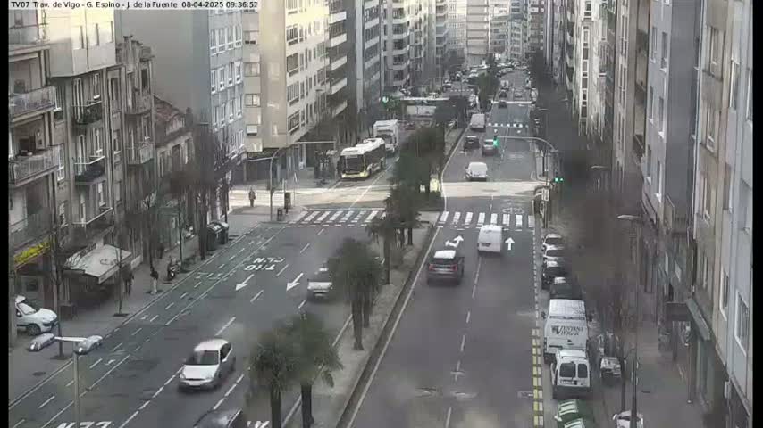 A city street scene shows several cars and vans driving on a multi-lane road, alongside buildings, with a bus stopped at a crosswalk.