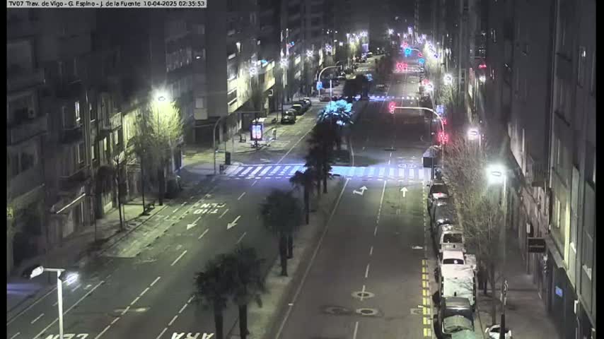 A nighttime street scene shows a multi-lane road with parked cars lining the sides, flanked by buildings with streetlights illuminating the area.