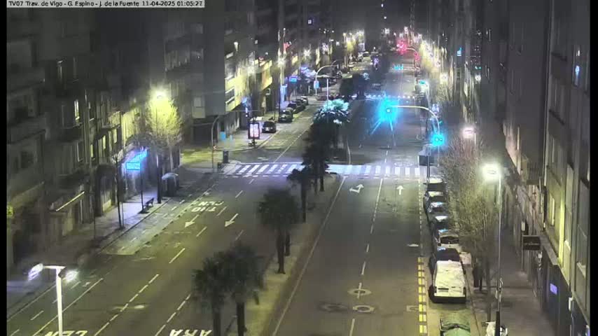 A nighttime, street-level view of a city street with cars parked along the sides and a few cars driving down the road, illuminated by streetlights.