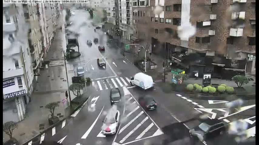 A rainy city street scene shows several cars driving on a wet road, with buildings lining both sides.
