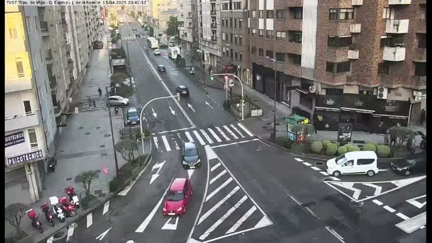 A city street intersection with several cars and pedestrians, under overcast daytime conditions.