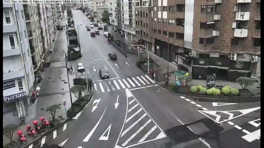 A wet city street intersection shows several cars and motorbikes, with buildings lining both sides of the road.