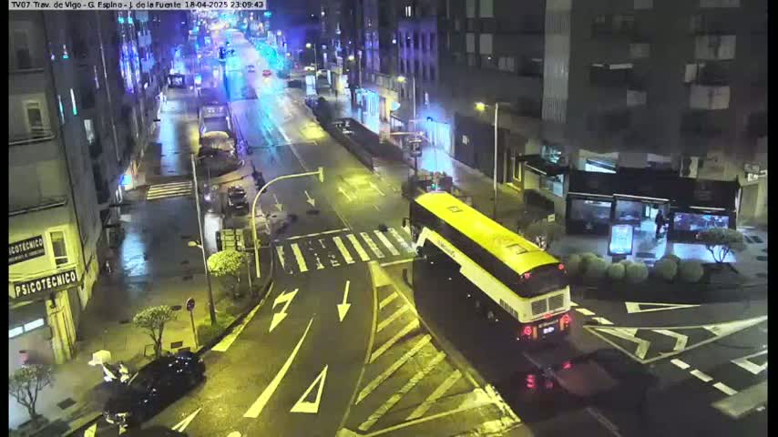 A nighttime street scene shows a wet road with a double-decker bus and several cars, surrounded by buildings.