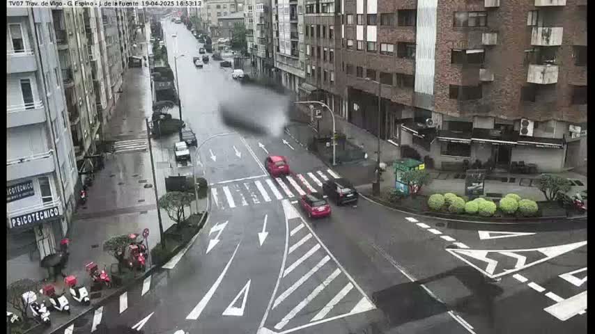 A rain-slicked city street intersection with several cars and motor scooters parked along the curb and several cars moving through the intersection.