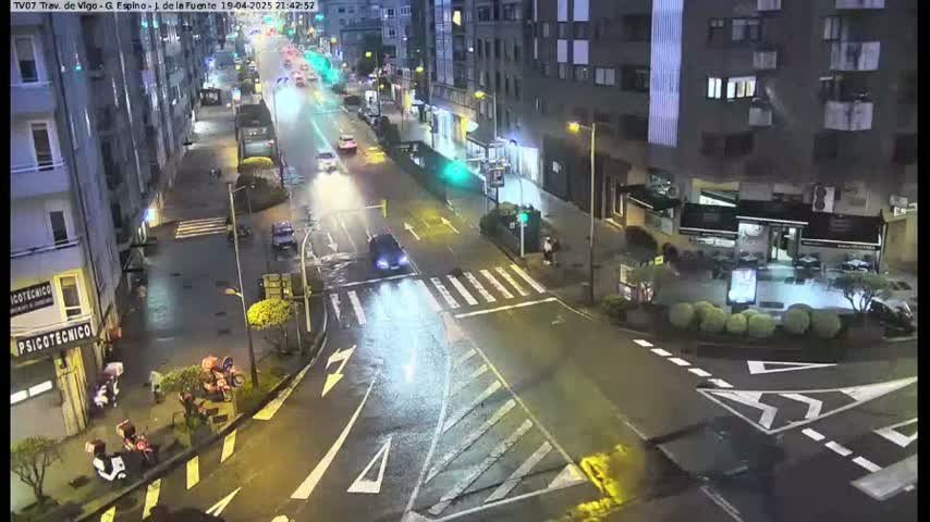 A wet, nighttime street intersection shows several vehicles and pedestrians in the city.