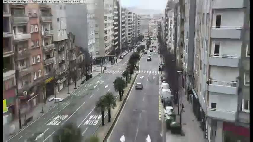 A mostly overcast day reveals a long city street lined with multi-story buildings, several cars are driving on the road, and some pedestrians are walking on the sidewalks.