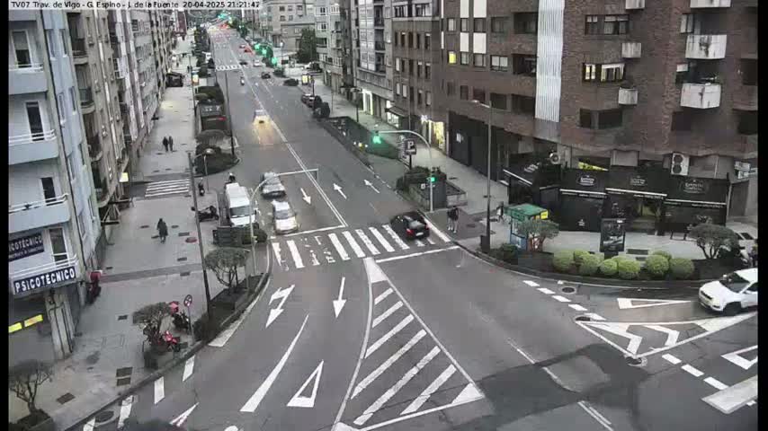 A street intersection with several cars and pedestrians, under overcast conditions.