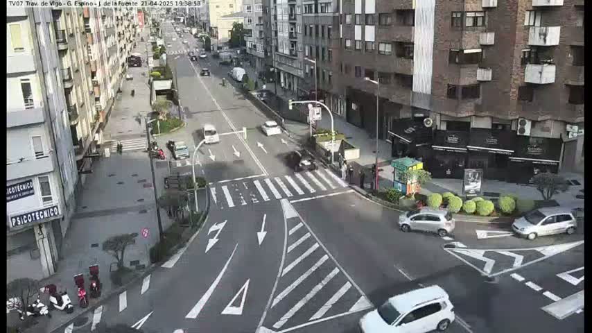 A city street intersection with several cars and pedestrians, under a bright, sunny sky.