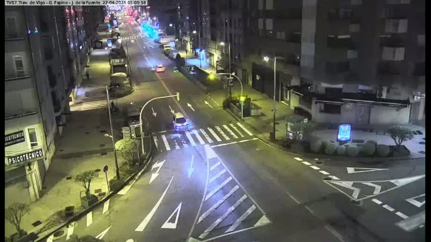 A nighttime, high-angle view of a mostly empty street intersection in a city, with a few cars visible and buildings lining the road.