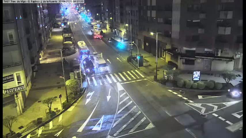 A nighttime, high-angle view of a city street intersection with several vehicles, including buses and cars, driving on roads lined with buildings.