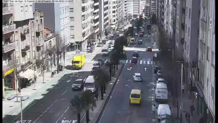 A sunny city street scene shows several cars and a van traveling in both directions, with pedestrians on the sidewalk and parked cars lining the street in front of multi-story buildings.