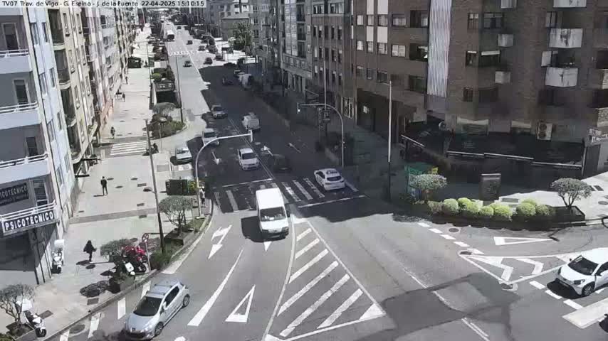 A sunny city intersection shows several cars and pedestrians navigating streets lined with multi-story buildings.
