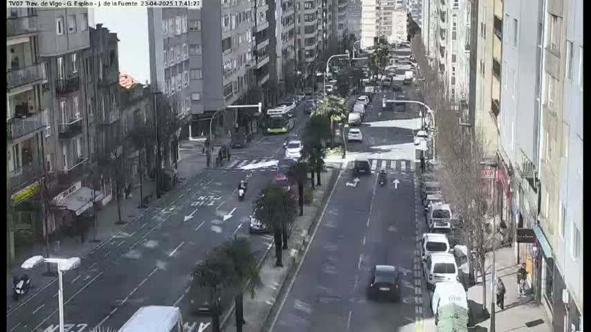 A city street scene on a sunny day shows several cars and a bus driving along a road lined with buildings and palm trees.