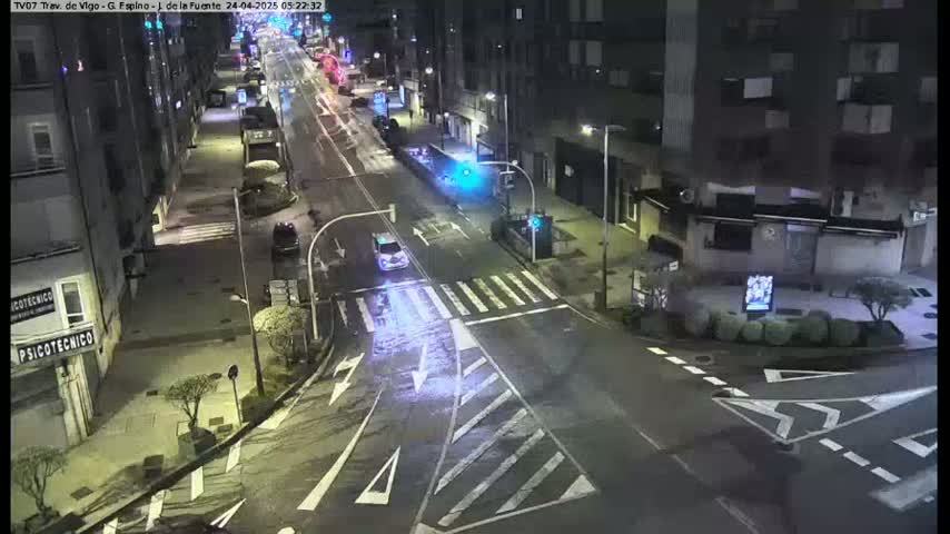 A nighttime view of a mostly empty city street intersection with a few cars, buildings lining the street, and pedestrian crosswalks.