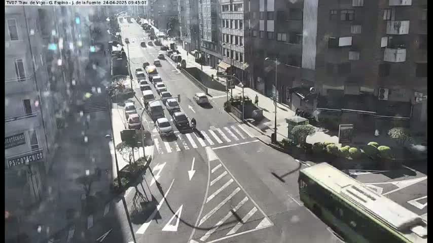 A sunny city street scene shows a multi-lane road with several cars and a bus, all moving in different directions, passing by multi-story buildings.