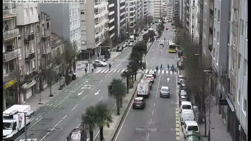 A city street scene on an overcast day shows several cars driving along a multi-lane road lined with buildings and palm trees.