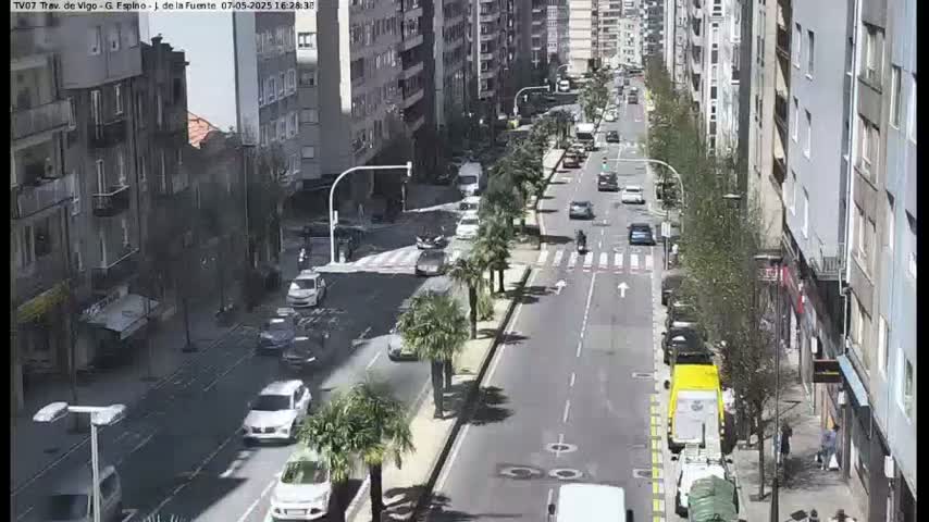 A sunny city street scene shows several cars driving in both directions past multi-story buildings and palm trees lining a median.