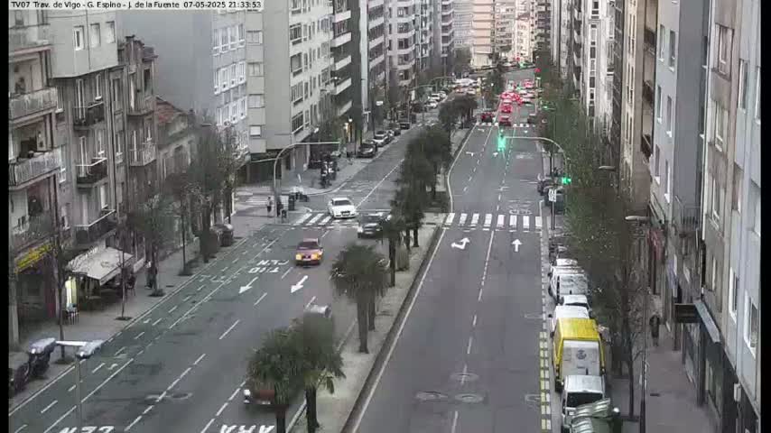 A city street scene at night shows several cars driving on a multi-lane road between tall buildings, lined with palm trees and parked vehicles.