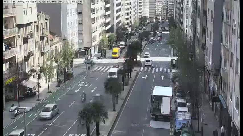 A sunny day shows a city street with cars and pedestrians, flanked by tall buildings and lined with trees.