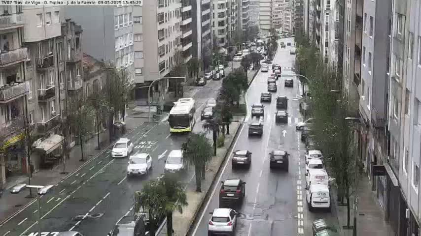 A rainy city street scene shows multiple cars driving in both directions past multi-story buildings, with a bus stopped at a bus stop.