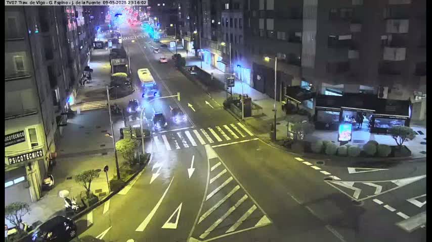 A nighttime street scene shows several cars and a bus at a crossroad intersection near some buildings.