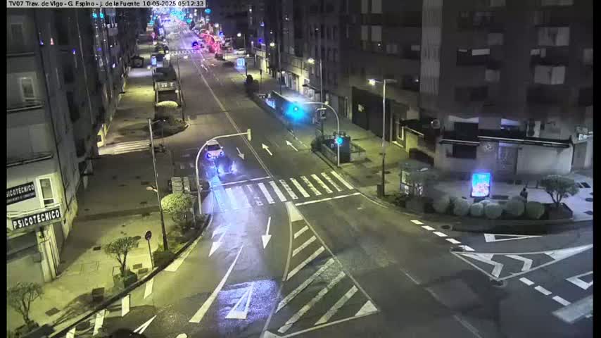 A nighttime street intersection with a few cars and buildings visible under partly cloudy skies.