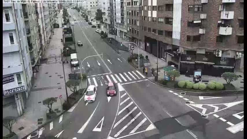A city street intersection with several cars, buildings on both sides of the street, and pedestrian crossings is shown under an overcast sky.