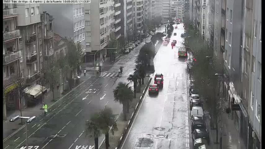 A rain-slicked city street lined with tall buildings has several cars driving on it, some pedestrians walking on the sidewalk, and palm trees planted along the median.