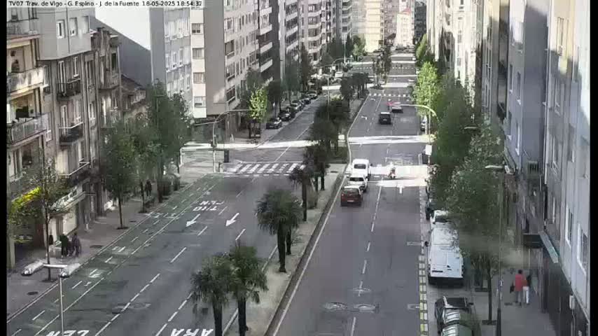 A city street scene on an overcast day shows several cars driving on a multi-lane road lined with buildings and palm trees.