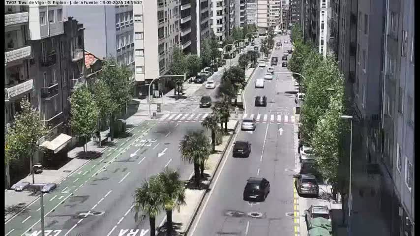 A city street scene on a partly sunny day shows several cars driving on a multi-lane road lined with buildings and palm trees.