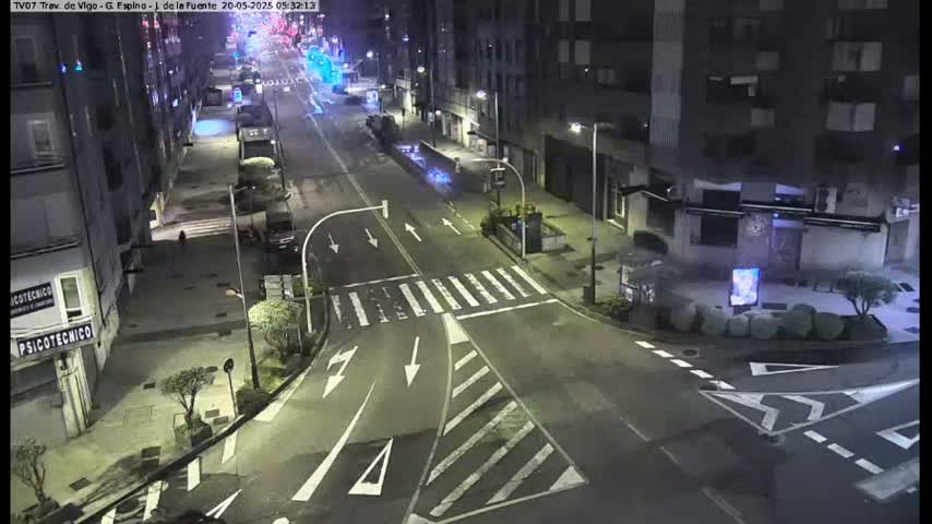 A nighttime street intersection with sparsely parked cars and few pedestrians, under artificial street lighting.