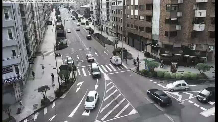 A street intersection in a city with several cars and pedestrians, under an overcast sky.