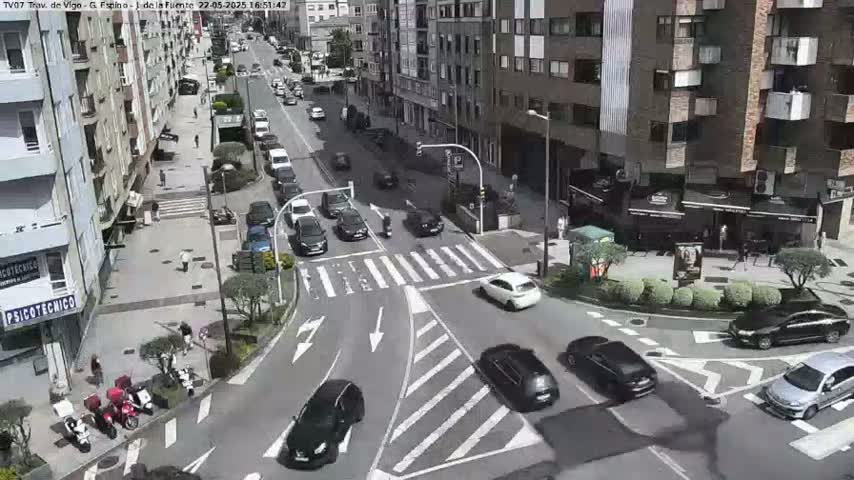 A sunny city street intersection shows several cars and pedestrians moving in and around a crosswalk.