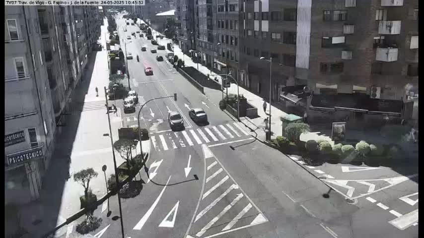 A sunny day shows a city street intersection with several cars and pedestrians, with multi-story buildings lining the street.