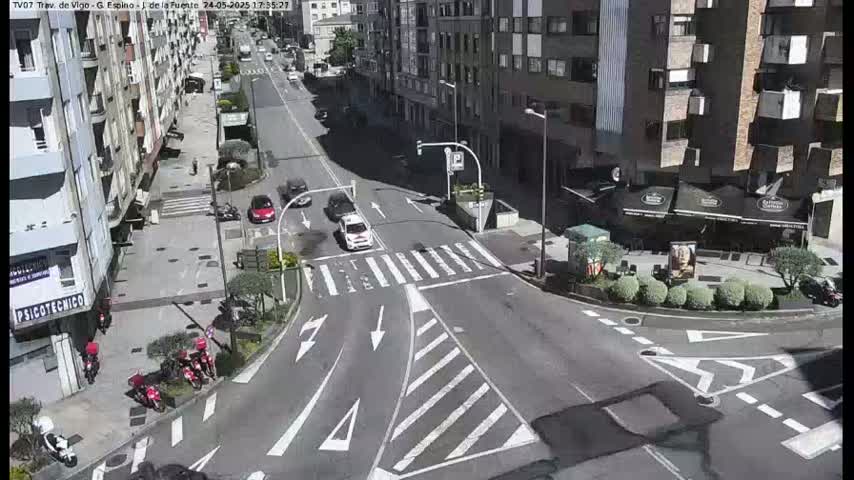A sunny day shows a city street intersection with several cars and motorcycles parked along the sides of the road and a few driving through the intersection.
