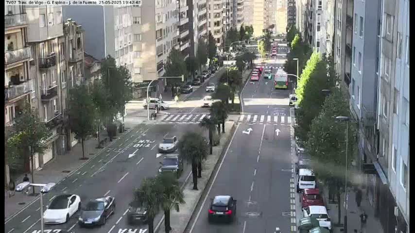 A sunny city street scene shows several cars driving along a multi-lane road lined with buildings and palm trees.