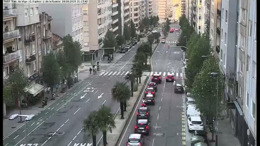 A city street with several cars driving in both directions, lined with tall buildings and palm trees on a partly cloudy day.