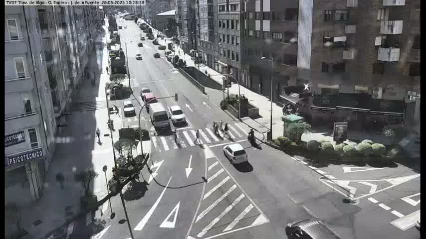 A sunny city street scene shows several cars and pedestrians at a crosswalk, with multistory buildings lining the street.