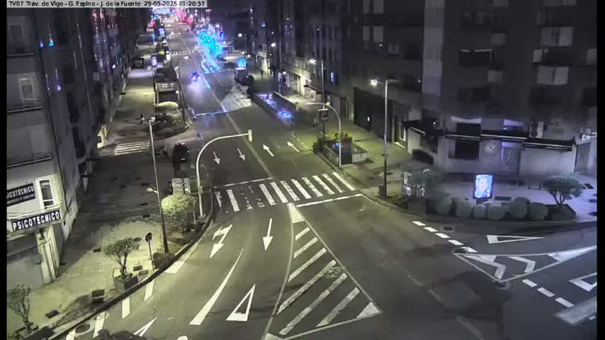 A nighttime street scene shows a mostly empty intersection with several cars, buildings lining both sides of the street, and some landscaping.