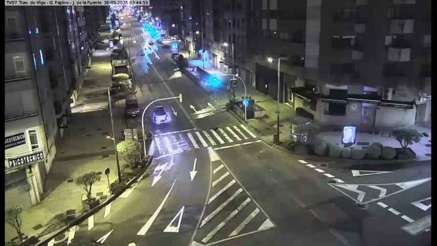 A nighttime, street-level view shows a mostly empty intersection with a few cars and buildings along the road.