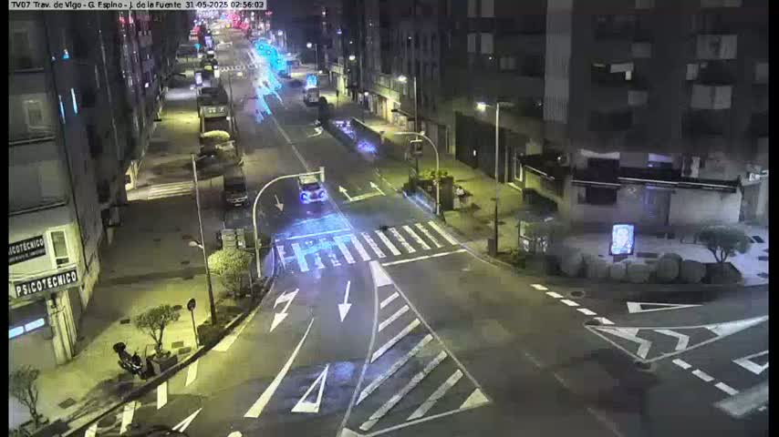 A nighttime street scene shows a mostly empty intersection with a few cars and a motorcycle, bordered by multi-story buildings.