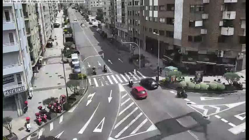 A city street intersection with several cars and motorcycles, surrounded by buildings, on a sunny day.